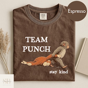 Pode incluir: Camiseta cor de café com as palavras "TEAM PUNCH" e "stay kind" impressas em branco. O gráfico mostra um macaco de desenho animado puxando outro macaco pela cauda. A camisa está dobrada.
