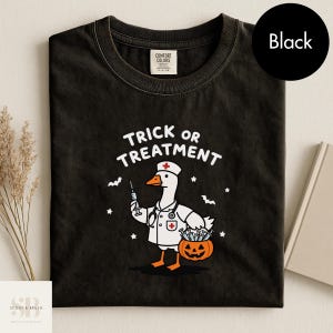 May include: A black t-shirt featuring a white graphic of a goose dressed as a nurse, holding a syringe and a jack-o'-lantern filled with syringes. The text above the goose reads "TRICK OR TREATMENT" in white letters, surrounded by small stars and bats.