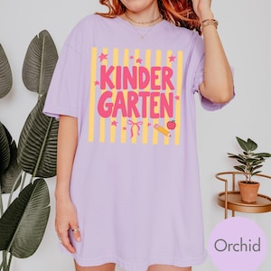May include: A light purple short-sleeve t-shirt featuring a graphic print with vertical yellow stripes and the pink text "KINDERGARTEN". The design includes small pink stars, a pink bow, a yellow pencil, and a red apple icon centered on the chest.