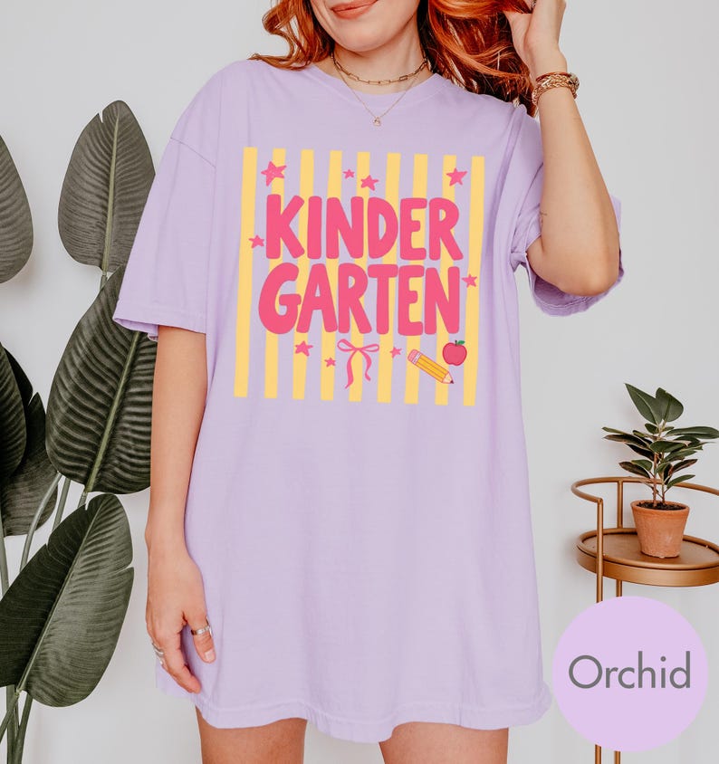 May include: A light purple short-sleeve t-shirt featuring a graphic print with vertical yellow stripes and the pink text "KINDERGARTEN". The design includes small pink stars, a pink bow, a yellow pencil, and a red apple icon centered on the chest.
