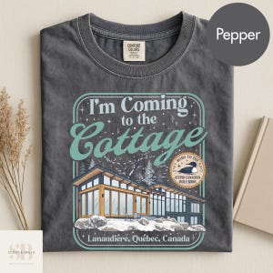 May include: Pepper-colored t-shirt with a graphic that reads "I'm Coming to the Cottage." The design features a modern cabin illustration with the text "Lanaudière, Québec, Canada."