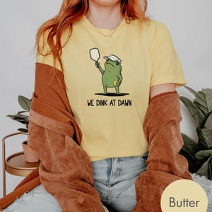 K&ouml;nnte beinhalten: Gelbes T-Shirt mit einem Cartoon-Frosch, der einen Pickleball-Schl&auml;ger h&auml;lt und ein Visier tr&auml;gt. Der Text "WE DINK AT DAWN" ist darunter gedruckt. Das Shirt wird mit blauen Jeans und einer braunen Jacke getragen.