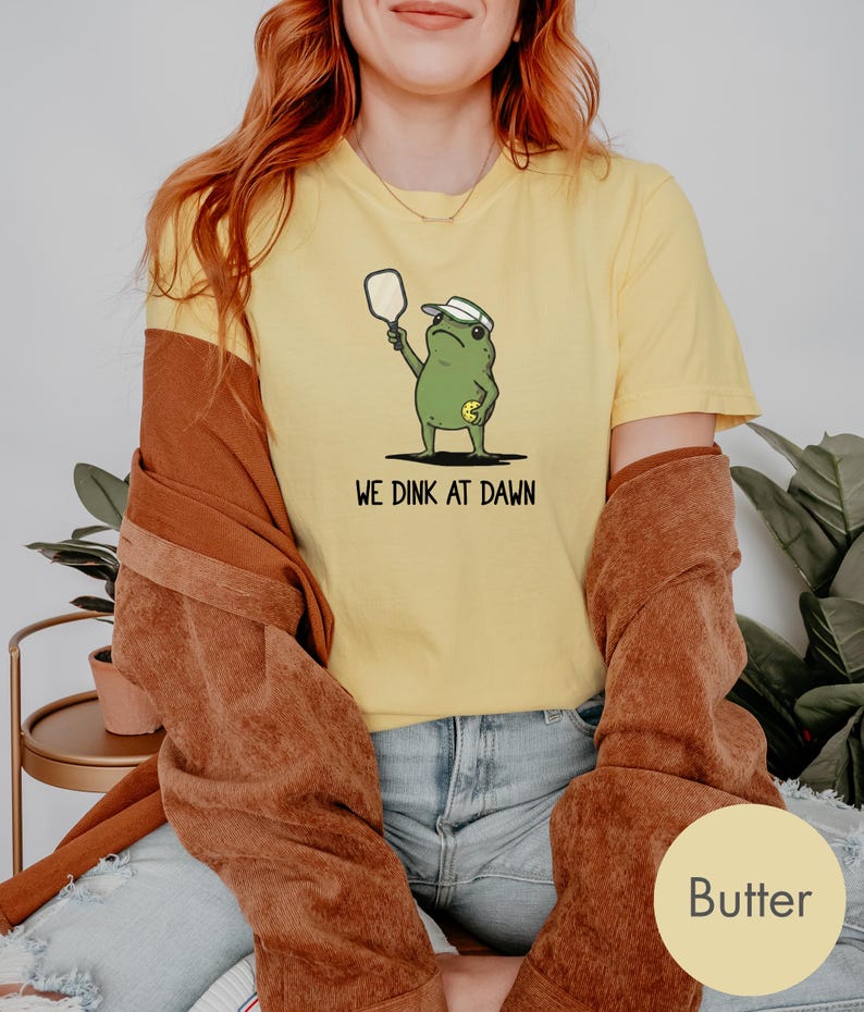 K&ouml;nnte beinhalten: Gelbes T-Shirt mit einem Cartoon-Frosch, der einen Pickleball-Schl&auml;ger h&auml;lt und ein Visier tr&auml;gt. Der Text "WE DINK AT DAWN" ist darunter gedruckt. Das Shirt wird mit blauen Jeans und einer braunen Jacke getragen.