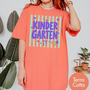 May include: A terra cotta t-shirt featuring a light green vertical striped square with the purple text KINDER GARTEN. The design includes purple stars, a bow, a yellow pencil, and a blue apple graphic centered on the front of the garment.