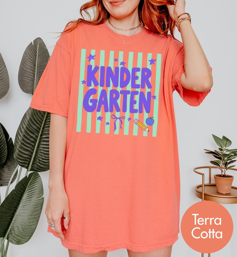 May include: A terra cotta t-shirt featuring a light green vertical striped square with the purple text KINDER GARTEN. The design includes purple stars, a bow, a yellow pencil, and a blue apple graphic centered on the front of the garment.