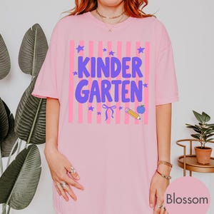 May include: A light pink t-shirt featuring a graphic with vertical pink stripes and the purple text "KINDERGARTEN". The design includes small purple stars, a purple bow, a yellow pencil, and a blue apple icon centered on the front of the shirt.
