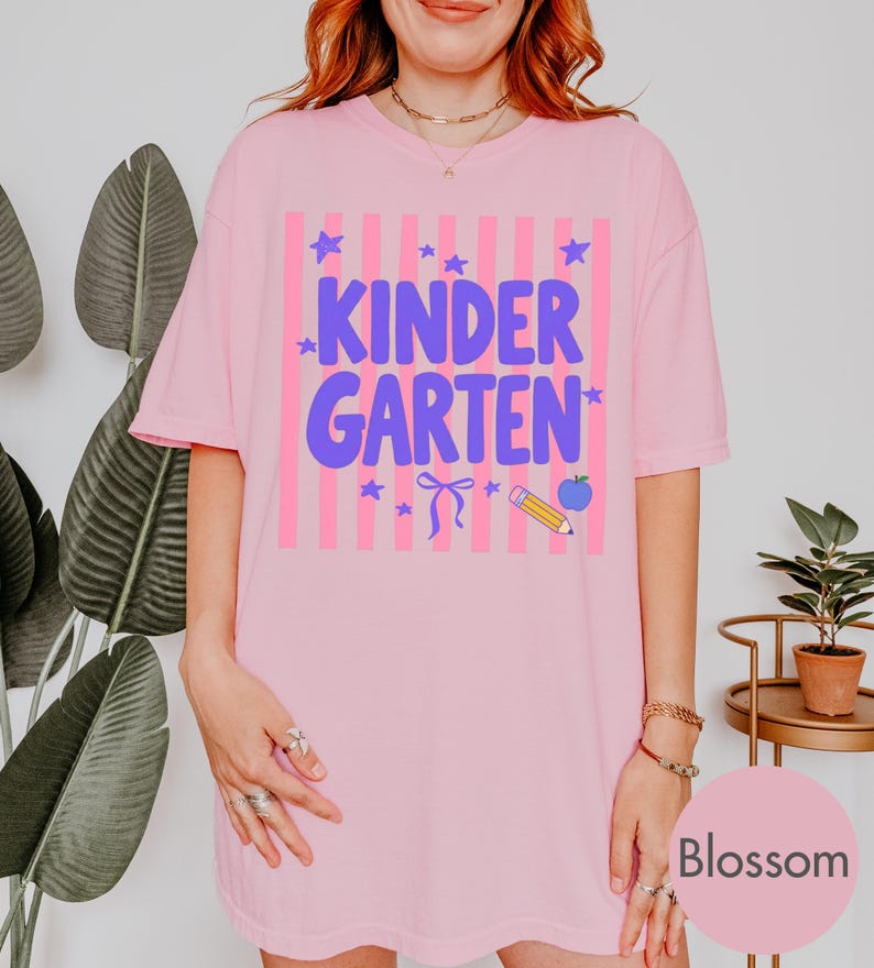 May include: A light pink t-shirt featuring a graphic with vertical pink stripes and the purple text "KINDERGARTEN". The design includes small purple stars, a purple bow, a yellow pencil, and a blue apple icon centered on the front of the shirt.