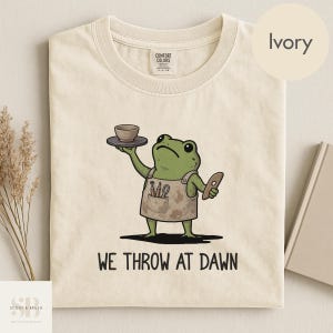 May include: An ivory t-shirt featuring a graphic of a green frog wearing a tan apron, holding a small bowl on a plate and a pottery tool. The frog is surrounded by the text WE THROW AT DAWN. The shirt has a Comfort Colors brand neck label.