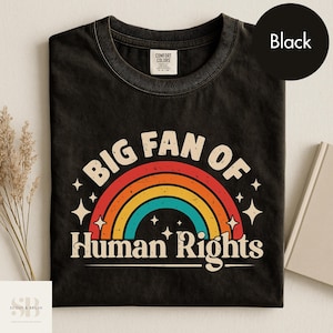 以下が含まれることがあります： 虹色のグラフィックと「BIG FAN OF HUMAN RIGHTS」の文字が入った黒いTシャツ。 デザインは星の付いた虹が特徴で、Comfort Colorsのシャツにプリントされています。