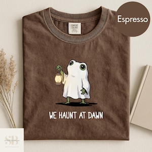 Peut inclure: T-shirt couleur expresso avec un motif de grenouille déguisée en fantôme tenant une lanterne. Le texte "WE HAUNT AT DAWN" est imprimé en dessous. Le t-shirt est en coton Comfort Colors.