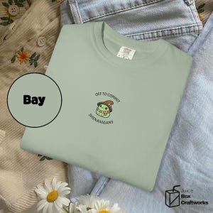 May include: A pale green t-shirt featuring the phrase "OFF TO COMMIT SHENANIGANS" and a cartoon frog wearing a hat. The shirt is folded on top of blue denim jeans and a beige corduroy shirt with floral embroidery.