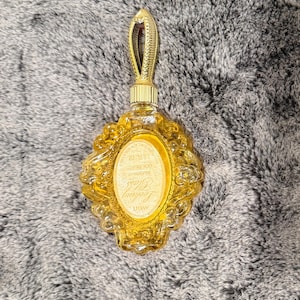 May include: A vintage, ornate perfume bottle in a golden hue. The bottle has a decorative handle and a detailed, oval-shaped body. The label reads "Golden Glass" and "1.5 fl oz".