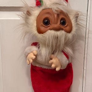 May include: A plush troll doll with a brown face and large dark eyes, wearing a red Santa hat with white faux fur trim and a small green holly accent. The doll has a long white faux fur beard and is attached to a red velvet Christmas stocking.
