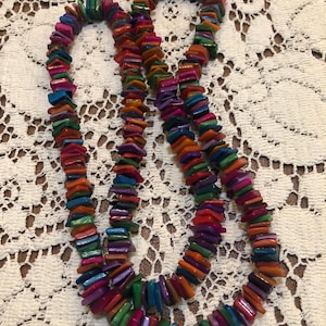 May include: A colorful necklace composed of small, rectangular beads in shades of blue, green, orange, purple, and red. The beads are strung together to form a long, layered necklace. The necklace is displayed on a white, lace-like surface.