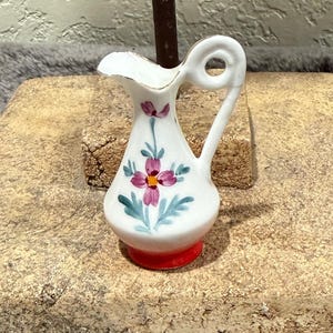 May include: A white ceramic mug with a black handle and the text "But First, Coffee" in a black, handwritten-style font. The mug is on a white surface.