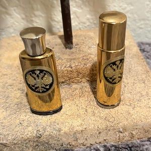 May include: Two cylindrical perfume bottles with a metallic gold finish. Each bottle features a circular black emblem on the front displaying a gold double-headed eagle. The left bottle has a silver-toned cap, while the right bottle has a matching gold cap.