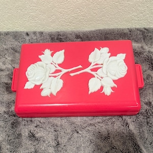 May include: A rectangular, bright pink container with a white floral design. The design features two white rose branches with leaves. The container has two small, red handles on the sides.