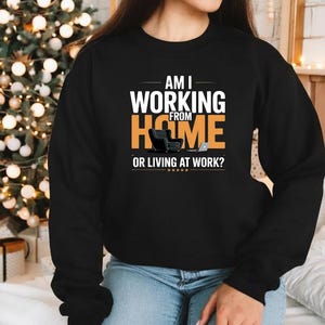 Peut inclure: Sweat-shirt noir avec l'inscription "AM I WORKING FROM HOME OR LIVING AT WORK?" en blanc et orange. Le motif comprend une petite chaise et un ordinateur portable. Un sapin de Noël décoré est en arrière-plan.