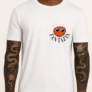 Puede incluir: Camiseta blanca con una cara de dibujos animados naranja y la palabra "Fantazia" en negro. El diseño está en el área del pecho izquierdo. La camiseta es de estilo cuello redondo.