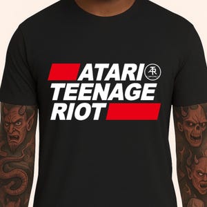 Könnte beinhalten: Schwarzes T-Shirt mit dem weißen Schriftzug "ATARI TEENAGE RIOT" und roten horizontalen Balken. Das Atari-Logo befindet sich in einem weißen Kreis. Ein Band-T-Shirt.