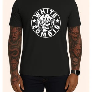 Puede incluir: Camiseta negra con un gráfico circular blanco con el texto "WHITE ZOMBIE" alrededor de una cara estilizada. La camiseta de manga corta está hecha de una tela oscura. La persona que lleva la camiseta tiene tatuajes en los brazos.