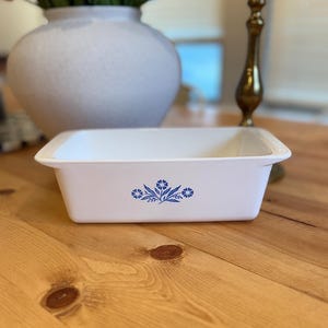 Puede incluir: Una fuente rectangular blanca para hornear con un diseño floral azul. La fuente tiene un borde ligeramente elevado y está sobre una superficie de madera. Un jarrón blanco con flores rosas y blancas está en el fondo.