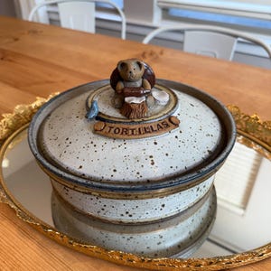 May include: A ceramic tortilla keeper with a speckled, off-white and brown finish. The lid features a bear figurine and the word "TORTILLAS". The keeper sits on a gold-rimmed, oval mirror.