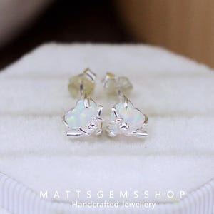 Może przedstawiać: Kolczyki sztyfty w kształcie serca z białymi opalami i srebrnymi oprawami. Kolczyki są prezentowane na białej powierzchni. Tekst "MATTSGEMSSHOP Handcrafted Jewellery" jest widoczny na dole.