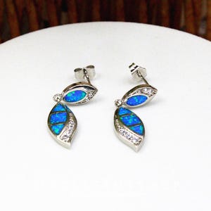 May include: A pair of silver-toned stud earrings with a leaf-shaped design. Each earring features a vibrant blue opal center, accented with clear crystals and a dangling opal leaf. The earrings are displayed on a white surface.
