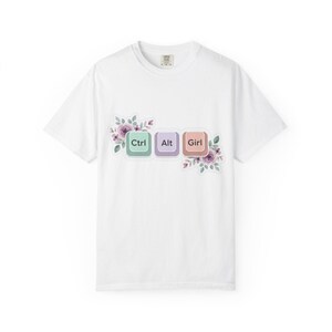 Puede incluir: Camiseta blanca con un diseño de tres teclas de teclado: Ctrl, Alt y Girl, en colores pastel. Elementos florales rodean las teclas. Prenda de vestir informal.