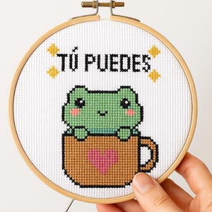 Puede incluir: Bordado de punto de cruz en un aro de madera. El diseño presenta una rana verde en una taza marrón con un corazón rosa y el texto "Tú Puedes" en negro. Acentos de estrellas amarillas completan el diseño.