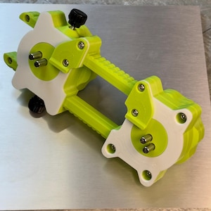 May include: A lime green and white mechanical device with black knobs and silver hardware. The device is positioned on a silver metal surface. The design features a series of interlocking components.