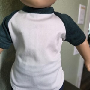 Puede incluir: Una mu&ntilde;eca viste una camiseta blanca de manga corta con mangas ragl&aacute;n de color verde oscuro y un cuello redondo a juego. La camiseta tiene el torso blanco liso y se combina con pantalones vaqueros azul oscuro, visibles en la parte inferior.