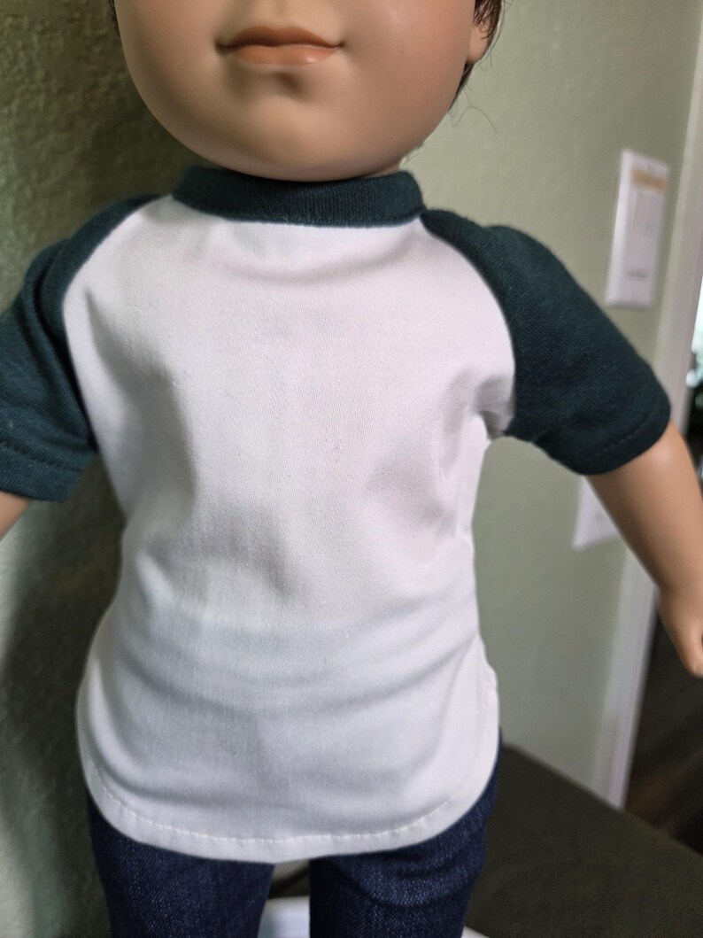 Puede incluir: Una mu&ntilde;eca viste una camiseta blanca de manga corta con mangas ragl&aacute;n de color verde oscuro y un cuello redondo a juego. La camiseta tiene el torso blanco liso y se combina con pantalones vaqueros azul oscuro, visibles en la parte inferior.