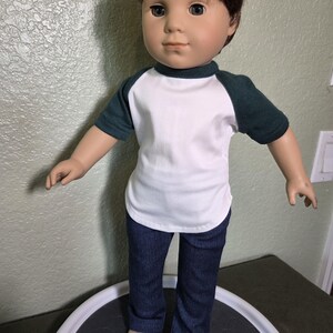 Puede incluir: Una mu&ntilde;eca con cabello casta&ntilde;o y ojos azules que viste una camiseta blanca con mangas ragl&aacute;n de color verde oscuro y pantalones de mezclilla azul oscuro. La mu&ntilde;eca tiene una expresi&oacute;n facial neutra y est&aacute; de pie con los brazos separados.