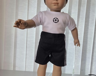 Puede incluir: Una muñeca de pelo y ojos castaños que viste una camiseta blanca de manga corta con un pequeño gráfico de un balón de fútbol en blanco y negro en el pecho, pantalones cortos deportivos negros con finas rayas blancas y zapatillas deportivas.