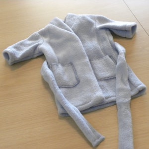 Op de afbeelding: Een lichtblauwe fleece badjas met twee voorzakken en een bijpassende riem. De badjas is gemaakt van een zacht, pluche materiaal. Het kledingstuk is waarschijnlijk voor een kind of een kleine pop.