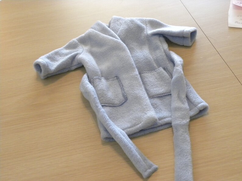 Op de afbeelding: Een lichtblauwe fleece badjas met twee voorzakken en een bijpassende riem. De badjas is gemaakt van een zacht, pluche materiaal. Het kledingstuk is waarschijnlijk voor een kind of een kleine pop.