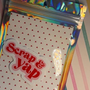 Op de afbeelding: Een doorzichtige, iriserende plastic zak met een witte kaart met een patroon van kleine rode hartjes. Een sticker met de woorden "Scrap & Yap" in rood script staat op de kaart. De achtergrond heeft een gestreept patroon van roze, blauw en wit.