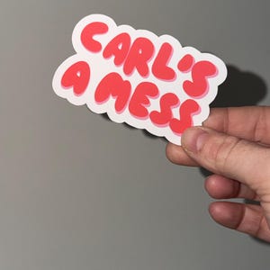 May include: A sticker with the text "CARL'S A MESS" in red bubble letters. The sticker has a white border and is held against a gray background.