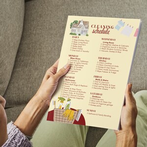 May include: A printed cleaning schedule with a house illustration at the top. The schedule is divided into daily, weekly, and weekend tasks. The text "Cleaning schedule" is at the top in a decorative font. The schedule is held by a person.