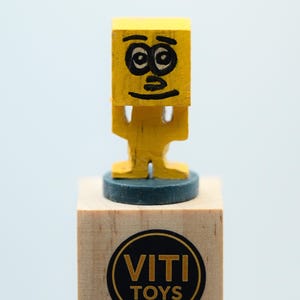 May include: A yellow wooden toy figure with a square head and a painted face, standing on a wooden block. The block has a circular sticker with the words "VITI TOYS". The figure has a simple, cartoonish design.