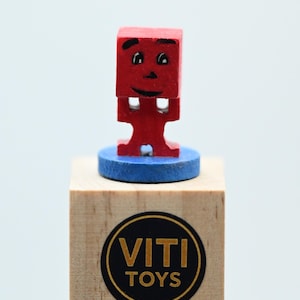 Pode incluir: Uma pequena figura de brinquedo de madeira vermelha com um rosto sorridente, em pé sobre uma base circular azul. A figura é montada em um bloco de madeira com o logotipo "VITI TOYS" em um círculo preto e dourado.
