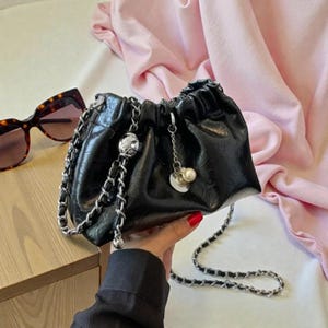 Puede incluir: Bolso de mano negro fruncido con una correa de cadena plateada y detalles. El bolso tiene un acabado brillante y texturizado y un pequeño dije con perlas. Gafas de sol visibles en el fondo.
