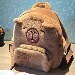 May include: A brown plush backpack shaped like a bear with a small front pocket. The backpack has a zipper closure and adjustable straps. The front pocket has the text "capy bara" in a star-shaped patch.