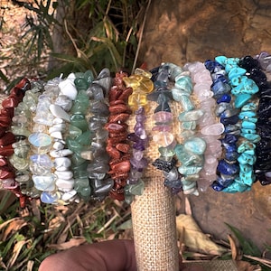 May include: A collection of gemstone chip bracelets in various colors, including red, green, yellow, pink, blue, and purple. The bracelets are displayed on a tan fabric-covered stand, showcasing the natural variations in the stones.