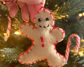 May include: A felt gingerbread man Christmas ornament with red stitching and bead accents. The ornament has a smiling face with black eyes and is suspended by a red and white string. Festive holiday decor.