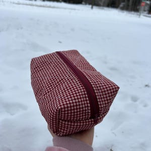 May include: A rectangular, quilted cosmetic bag with a red and white gingham pattern. The bag has a burgundy zipper closure and is held up against a snowy background. The bag appears to be made of fabric.