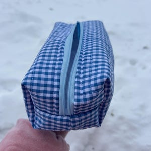 Könnte beinhalten: Ein blau-weiß kariertes Etui mit Reißverschluss. Das Etui ist rechteckig mit einem hellblauen Reißverschluss und einem Steppmuster. Der Hintergrund ist eine verschneite Landschaft.