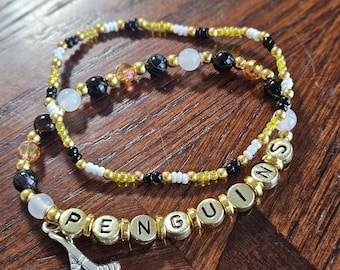 以下が含まれることがあります： 「PENGUINS」の文字が金色のビーズで綴られた、3つのビーズブレスレット。ブレスレットは、黒、白、金、透明のビーズを使用。シルバーのホッケースティックチャームが1つのブレスレットにぶら下がっています。